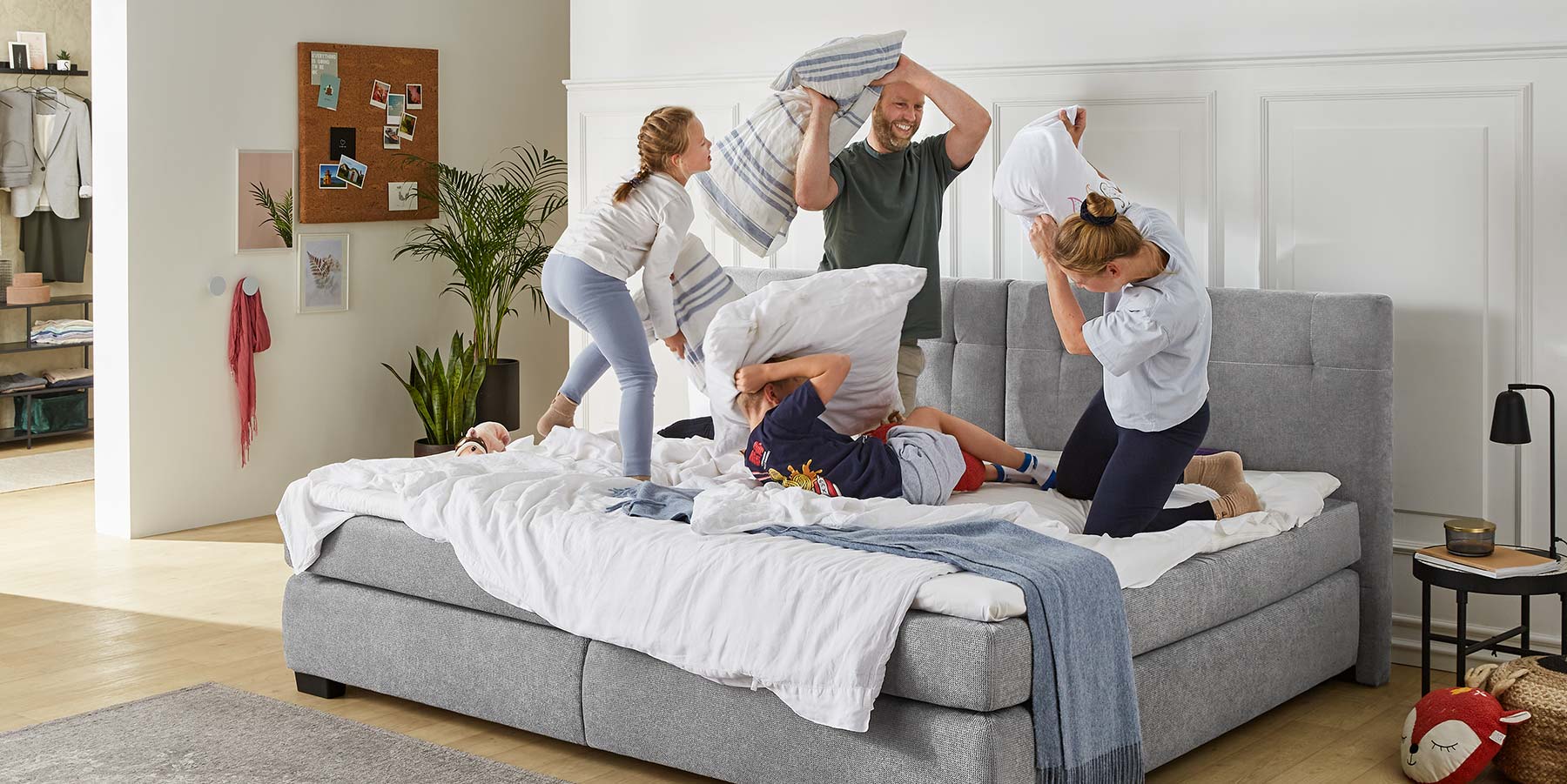 Zeigt eine glückliche Familie die mit ihren Kindern eine Kissenschlacht auf einem gemütlichen Boxspringbett macht.